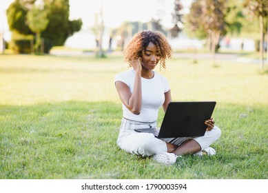 Young Attractive Dark-skinned College Student Wearing Tank Top And Jeans Sitting On The Lawn At Campus On Sunny Day, Working On Her Thesis Using Laptop Computer, Looking Busy And Concentrated