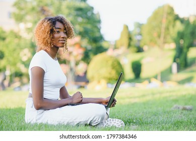 Young Attractive Dark-skinned College Student Wearing Tank Top And Jeans Sitting On The Lawn At Campus On Sunny Day, Working On Her Thesis Using Laptop Computer, Looking Busy And Concentrated