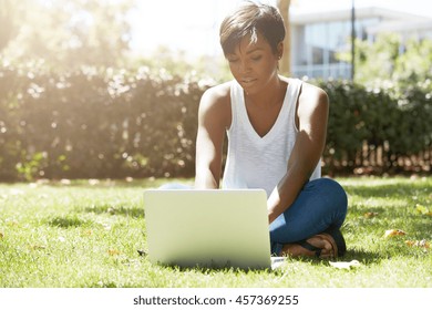 Young Attractive Dark-skinned College Student Wearing Tank Top And Jeans Sitting On The Lawn At Campus On Sunny Day, Working On Her Thesis Using Laptop Computer, Looking Busy And Concentrated