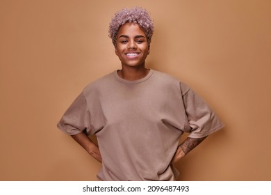 Young attractive ethnic African American woman curly hairstyle smiles broadly and looks at camera holds hands on belt posing standing in beige oversized t-shirt on brown studio background - Shutterstock ID 2096487493