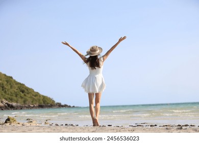 Young attractive ethnic Thai millennial woman posing at suny spreading arms to sides and various postures dressed in white casual clothes stands on the ocean with sandy beach. Love for travel - Powered by Shutterstock