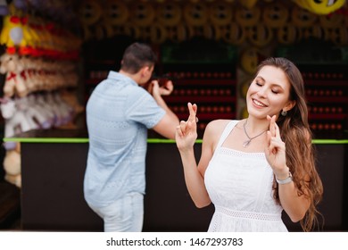 Young Attractive European Couple In Amusement Park. Male And Female On Romantic Date. Guy Shoots A Gun Into Shooting Range To Win A Toy For The Girl. She Crossed Her Fingers To Get A Prize