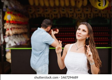 Young Attractive European Couple In Amusement Park. Male And Female On Romantic Date. Guy Shoots A Gun Into Shooting Range To Win A Toy For The Girl. She Crossed Her Fingers To Get A Prize