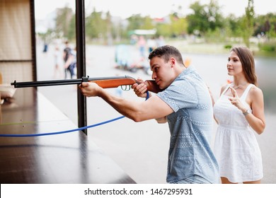 Young Attractive European Couple In Amusement Park. Male And Female On Romantic Date. Guy Shoots A Gun Into Shooting Range To Win A Toy For The Girl. She Crossed Her Fingers To Get A Prize