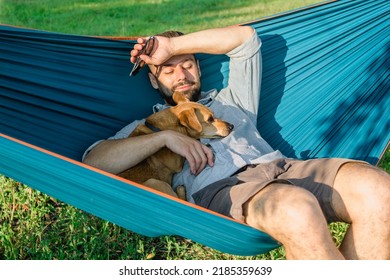 Young attractive European man is dozing in hammock with his cute little dog. Sunny weekend in summer park. - Powered by Shutterstock