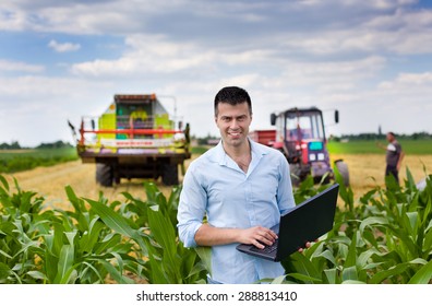 Young attractive farmer with laptop standing in corn field, tractor and combine harvester working in wheat field in background