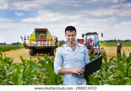 Young attractive farmer with laptop standing in corn field, tractor and combine harvester working in wheat field in background