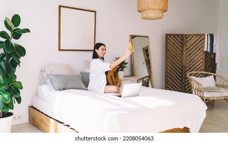 Young attractive female blogger smiles and posing for smartphone camera. Brunette woman vlogger recording video on mobile phone while sitting with guitar in bedroom interior - Shutterstock ID 2208054063