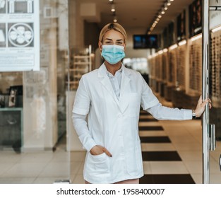 Young and attractive female optometrist with face protective mask standing at open optical store doors and looking outside. She is confident and serious. Covid-19 open for business concept.