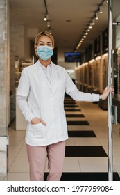 Young And Attractive Female Optometrist With Face Protective Mask Standing At Open Optical Store Doors And Looking Outside. She Is Confident And Serious. Covid-19 Open For Business Concept.