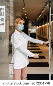 Young And Attractive Female Optometrist With Face Protective Mask Standing At Open Optical Store Doors And Looking Outside. She Is Confident And Serious. Covid-19 Open For Business Concept.