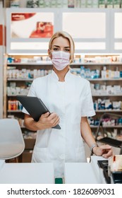 Young And Attractive Female Pharmacist With Face Protective Mask Working In Drugstore. She Is Confident And Serious. Covid-19 Open For Business Concept.