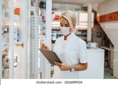 Young And Attractive Female Pharmacist With Face Protective Mask Working In Drugstore. She Is Confident And Serious. Covid-19 Open For Business Concept.