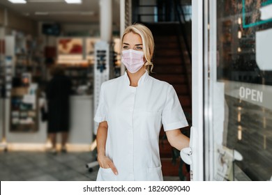 Young And Attractive Female Pharmacist With Face Protective Mask Standing At Open Drugstore Doors And Looking Outside. She Is Confident And Serious. Covid-19 Open For Business Concept.