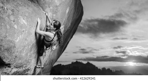 Young Attractive Female Rock Climber Climbing Challenging Route On Steep Rock Wall Against Scenic Sunset Background. Summer Time. Climbing Equipment. Black And White
