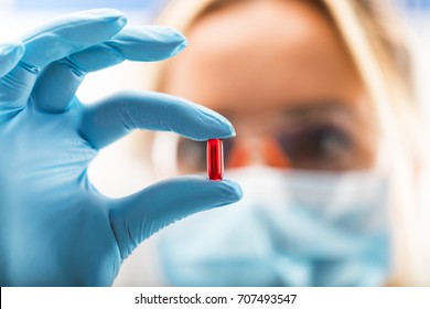 Junge attraktive Wissenschaftlerin mit Schutzbrille und Maske, die eine rote, transparente Tablette mit Fingern in Handschuhen im pharmazeutischen Forschungslabor hält