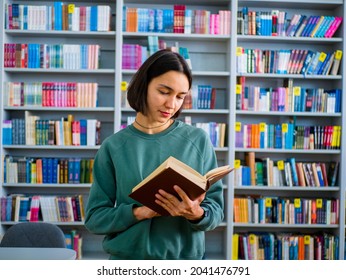 Young attractive female student reads a book preparing for an exam while against the backdrop of bookshelves.Education concept.