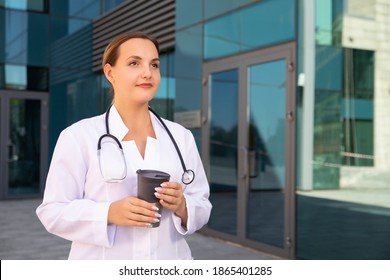 Young attractive female surgeon holding hot cup of coffee in her hands, wearing medical white coat, hanging stethoscope around her neck, looking rested. Medicine, healthcare and urban concept. - Powered by Shutterstock