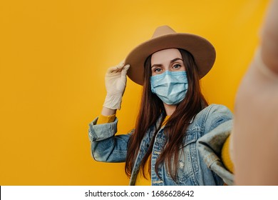 Young attractive female traveler in hat and gloves makes selfie against yellow background, dressed in denim jacket, wears protective medical mask to protect herself from coronavirus. Pandemic concept - Powered by Shutterstock