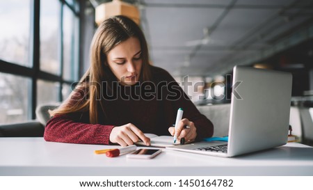 Young attractive female writer scribing notes to textbook prepare for exam, girl doing homework sitting desktop with modern laptop computer, woman making plan for tomorrow and creating  to do list