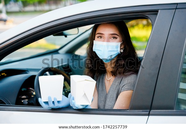 Young attractive girl in car in medical gloves and\
mask holds wok in box udon noodles with tempuru, shrimps, in hands\
and smiles. Udon noodles in white box delivery. 2 Wok box udon\
advertise 1+1.