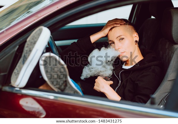 Young and attractive girl\
with a shaved head, vaping inside her car during a break on the\
trip.