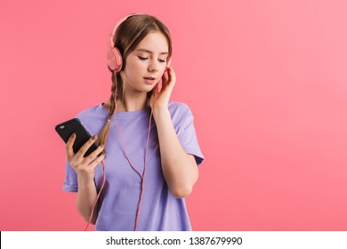 Young attractive girl with two braids in lilac t shirt listening music in headphones with cellphone in hand dreamily looking aside over pink background - Shutterstock ID 1387679990