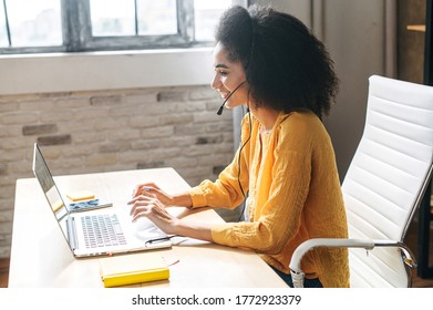 Young attractive girl uses headset for talk online while working in the light modern office. Side view of an ethnic woman with an afro hairstyle in the office