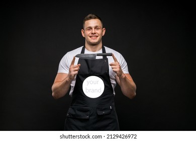 Young Attractive Guy With Big Muscles, Stylish Hairstyle In A White T-shirt And Black Apron With A Place For A Logo, Smiling Holding A Kitchen Knife With Both Hands On A Black Background