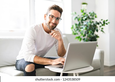 A Young attractive guy is browsing at his laptop, sitting at home on the cozy beige sofa at home, wearing casual outfit - Shutterstock ID 1243538509