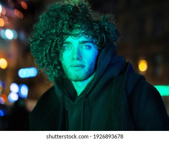 Young, Attractive, Handsome Curly Boy Poses On A Green Neon Background. Street, Urban Modeling, Portrait, Fashion Photo. Cyberpunk Style Photography With High Fashion Model. Green Neon Light.