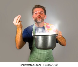 Young Attractive Happy And Proud Home Cook Man Cooking Soup Holding Kitchen Pot Smelling The Meal Aroma Gesturing Delighted And Satisfied On Isolated Background In Domestic Lifestyle