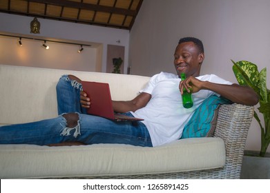 Young Attractive And Happy Successful Black African American Man Networking With Laptop Computer At Living Room Couch Smiling Cheerful Drinking Beer Bottle  In Internet Business Success Concept 