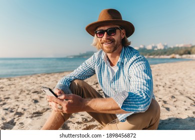 young attractive hipster man sitting on beach by sea on summer vacation, boho style outfit, holding using internet on smartphone, dressed in shirt, sunglasses and hat - Powered by Shutterstock