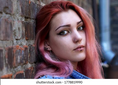 Young attractive hipster woman with pink hair and nose and lips piercing sitting against brick wall.