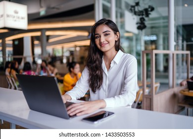 A Young And Attractive Indian Asian Woman Small Business Entrepreneur Smiles As She Works On Her Laptop In A Cafe During The Day. She Is Wearing A Business Casual Outfit And Is Hopeful And Confident. 