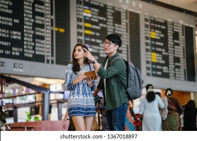A young, attractive interracial Asian couple are finding their way to their flight in an airport. The Korean man is pointing the way to his Indian girlfriend in front of the flight information display