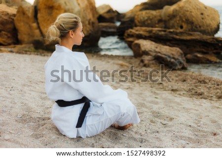 Young attractive karate woman in white kimono with a black belt meditation on wild beach with stones