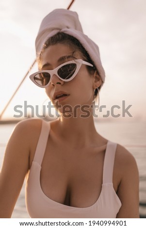 Young attractive lady with towel wrapped hair, in modern white swimsuit, round earrings and sunglasses resting at sea during pink sunset