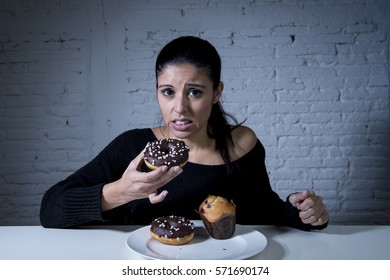 Young Attractive Latin Woman Sitting At Table Feeling Guilty Forgetting Diet Eating Dish Full Of Junk Sugary Unhealthy Food Such As Chocolate Donut And Muffin Cake In Sugar Addiction