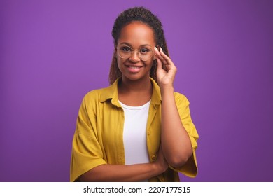 Young attractive long haired African American woman millennial adjusts glasses and smiling looks at camera posing in yellow casual shirt stands on purple studio background. Ethnic girl - Powered by Shutterstock