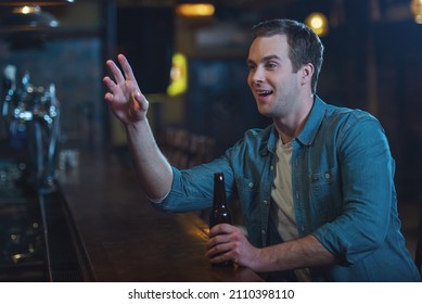 Young attractive man in casual clothes is smiling, holding a bottle of beer and making order while sitting at bar counter in pub - Powered by Shutterstock