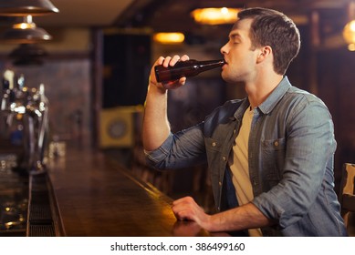 Young Attractive Man In Casual Clothes Is Drinking Beer While Sitting At Bar Counter In Pub