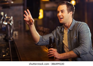 Young Attractive Man In Casual Clothes Is Smiling, Holding A Bottle Of Beer And Making Order While Sitting At Bar Counter In Pub