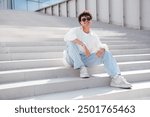 Young attractive man with curly hair hairstyle. Smiling  handsome male in casual stylish clothes posing in the street it sunny day. Cheerful and happy model outdoors, sits at stairs