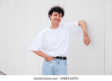 Young attractive man with curly hair hairstyle. Smiling  handsome male in casual stylish jeans and t-shirt clothes posing in the street it sunny day. Cheerful and happy model outdoors, near wall - Powered by Shutterstock