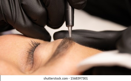 Young attractive man eye close-up with the master hand in black gloves and permanent makeup machine with a needle.