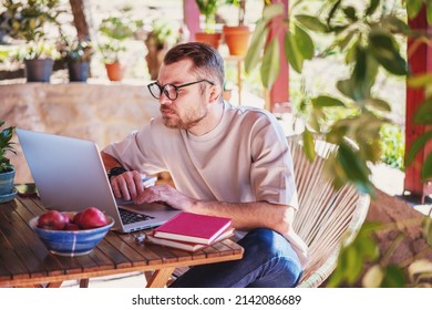 Young Attractive Man In Glasses Freelancer Working From Home Using Laptop While Sitting On The Terrace Of A Country House On A Summer Day