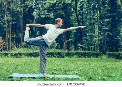 Young attractive man in a gray T-shirt and sweatpants doing yoga  pose in the park. Keeps balance on one leg