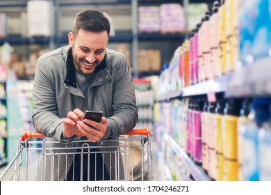 Young Attractive Man In Grey Coat Do Shopping In The Supermarket And Typing Phone